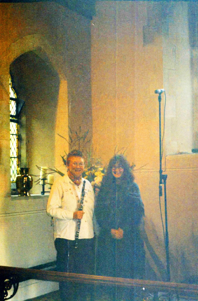 Two individuals stand together in a chapel, one holding a flute, surrounded by floral arrangements.